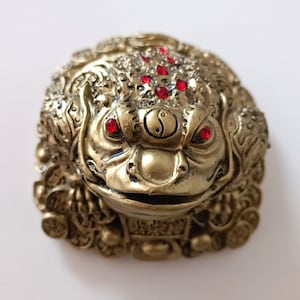 Puede incluir: Una figura de rana dorada de la fortuna con ojos de piedras preciosas rojas y un símbolo yin-yang en la frente. La rana está sentada sobre una pila de monedas de oro.