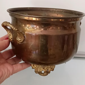 May include: A brass planter with ornate handles and feet. The planter is a deep copper color and has a smooth, shiny finish.