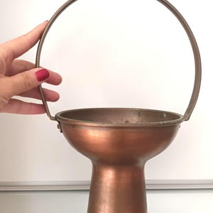 Puede incluir: Un cuenco de cobre con asa, el cuenco está sobre una base de pedestal.