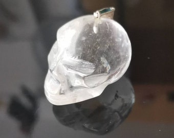 May include: A clear crystal skull pendant with a silver bail. The skull is carved with intricate details and has a smooth, polished finish.