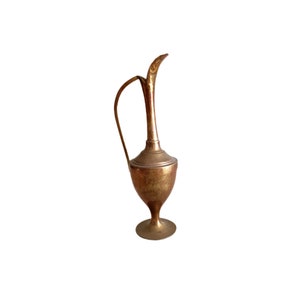 May include: A gold-toned brass pitcher with a curved spout and a round base. The pitcher has a worn, antique look.