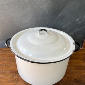 May include: A white enamelware pot with a lid. The pot has black trim around the rim and handles. The lid has a black handle. The pot is sitting on a wooden surface.