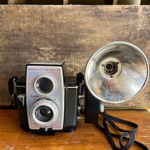 May include: A black Kodak Brownie Reflex 20 camera with a silver front panel and a large, circular, metallic flash attachment mounted on the side. The camera features a dual-lens system and a black fabric neck strap attached to the sides.