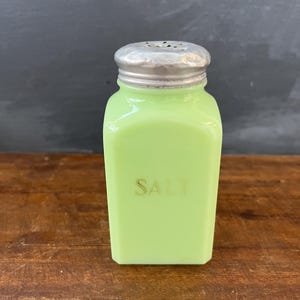 May include: A vintage green glass salt shaker with a silver lid and the word "SALT" printed on the front.
