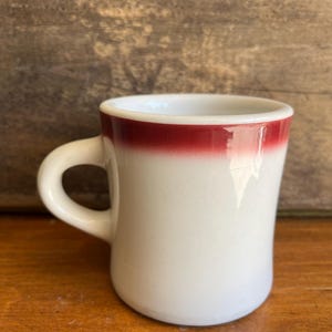 May include: A white ceramic coffee mug with a red rim and handle. The mug has a classic diner style and is set against a wooden background. The mug is empty.