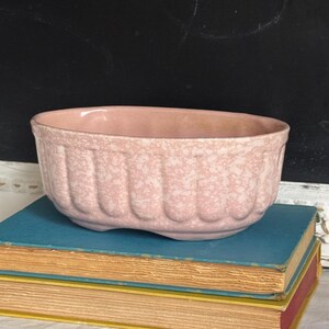May include: An oval ceramic planter with a light pink and white mottled glaze finish. The exterior features a vertical ribbed or fluted pattern along the sides, and the base is supported by two small, integrated feet for stability.