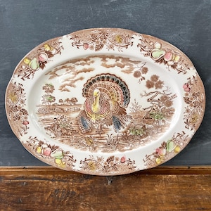 May include: Oval ceramic serving platter with a turkey illustration in the center. The platter has a brown border with floral and fruit designs. The turkey is depicted with colorful feathers, standing in a landscape scene.