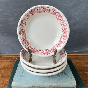 May include: A stack of four white ceramic saucers featuring a red scrollwork pattern along the rim. The top saucer is held upright by a small brass stand, displaying the intricate red floral-inspired filigree design against the white surface.