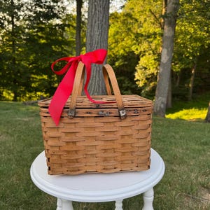 May include: A rectangular, woven wicker picnic basket with a red ribbon tied to the handle. The basket is brown and sits on a white, round table. The background features green grass and trees.