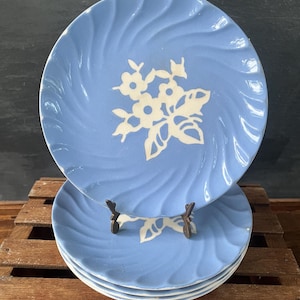 May include: A stack of light blue ceramic plates with a white floral design in the center. The plates have a swirled texture around the edges. The plates are displayed on a wooden surface.