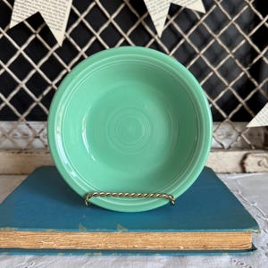 May include: A light green ceramic bowl with concentric circles in the center. The bowl is displayed on a gold-colored metal stand, resting on a blue book with aged pages. The background features a black grid and paper banner.