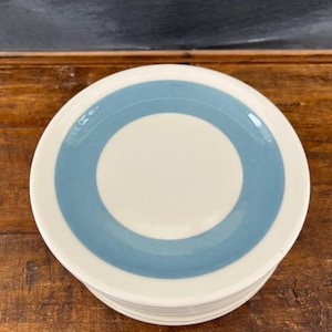 May include: A stack of white ceramic plates with a light blue ring design. The plates have a simple, modern aesthetic, suitable for everyday use. The plates are stacked on a wooden surface.
