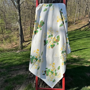 May include: A white tablecloth with a floral pattern of yellow and green flowers and leaves. The tablecloth is draped over a red wooden ladder, with a natural outdoor background.