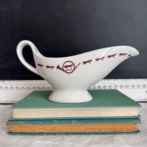 May include: White gravy boat with a handle and a flared spout. The boat is decorated with a series of maroon fox silhouettes and a hunting horn emblem. The gravy boat is resting on a stack of vintage books with green and blue covers.