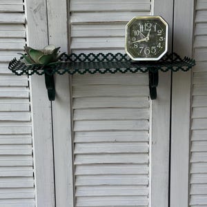 May include: A dark green metal shelf with a decorative edge, holding a green succulent and a vintage black and silver clock. The clock has a white face with black numbers. The shelf is mounted on a white shutter background.