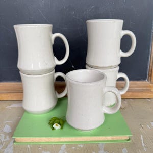 May include: A collection of off-white ceramic mugs with rounded handles. The mugs are stacked and arranged on a green book, with a small white flower and green leaves. The background is a dark chalkboard.