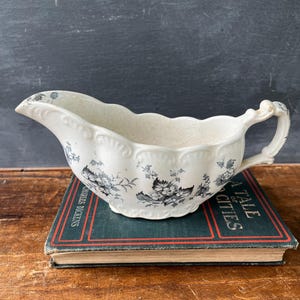 May include: A cream-colored ceramic gravy boat featuring a black floral transferware pattern, a scalloped rim, and an ornate handle. The vessel has a curved spout and sits atop a green hardcover book titled A Tale of Two Cities by Charles Dickens.