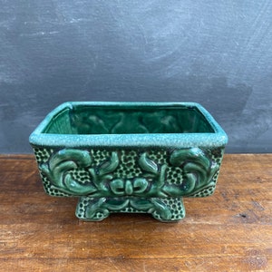 May include: A green ceramic planter with an ornate design and four legs. The planter is made of glazed ceramic and has a square shape.