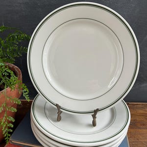 May include: A stack of white ceramic plates with a green border. The plates are stacked on top of each other, with one plate on a decorative metal stand. The plates are on a wooden surface.