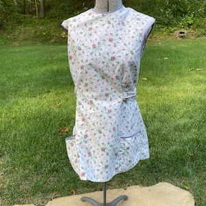May include: A white quilted apron with a floral print of pink roses. The apron has two pockets on the front and a tie at the waist.