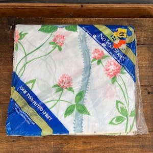 May include: A white twin fitted sheet with a floral pattern of pink and green clover flowers. The sheet is packaged in plastic and labeled "No Iron Sheet" and "Made of 65%  Dacron®  Polyester and 15% Cotton (Cotton/Co  Made in U.S.A. by Dan River Inc. Danville, Virginia 24141"