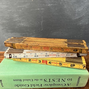 May include: Three vintage folding rulers stacked on a green book. The rulers are made of wood and metal, with inch markings. The book has the title "A Complete Field Guide to NESTS in the United States."