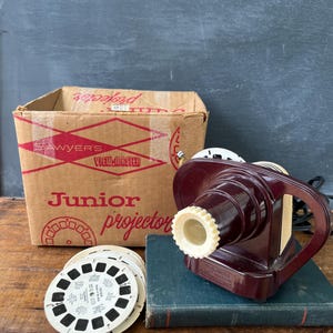 May include: A vintage Sawyer's View-Master Junior projector in a deep maroon and cream plastic housing, accompanied by its original cardboard box with red text and graphics, and a stack of circular white film reels for viewing images.