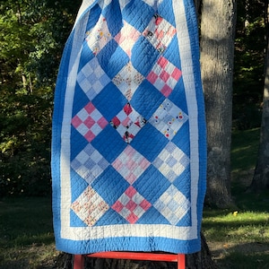 May include: A rectangular quilt featuring a diamond-patterned design with alternating solid blue squares and various multi-colored patterned squares. The quilt is bordered by a white band and finished with a solid blue outer edge, all featuring visible stitching.