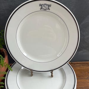 May include: White ceramic plates with a black rim and a thin black line inside the rim. The plates have a decorative emblem in the upper center. The plates are stacked on top of each other.