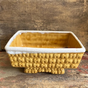 May include: A rectangular ceramic planter with a textured, golden-yellow exterior resembling corn kernels. The rim features a thick, white, speckled glaze that drips slightly down the sides. The vessel stands on a small, integrated pedestal base.