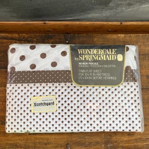 May include: A white twin flat sheet with brown polka dots. The sheet is packaged in plastic and has a label that says "Wondercale by Springmaid" and "No-Iron Percale 50% Kodel Polyester 50% Cotton". The label also says "Twin Flat Sheet for 39x76 in. Mattress (72x104 in. before hemming)". The sheet is treated with Scotchgard.