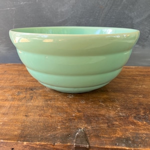 May include: A light green ceramic mixing bowl. The bowl has a rounded shape with a slightly flared rim and horizontal ridges around the exterior. The bowl is sitting on a wooden surface.