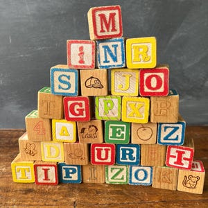 May include: A pyramid stack of wooden alphabet blocks featuring various letters, numbers, and illustrations including a bee, a baseball cap, a horse, a ball of yarn, an apple, and a mouse. The blocks are painted in red, blue, yellow, and green.