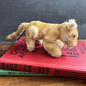May include: A vintage plush lion cub toy in tan and beige, with a detailed mane and tail. The toy is posed on top of a red book with the title "CHILD-LIFE ARITHMETICS". The lion has small red eyes and a detailed face.