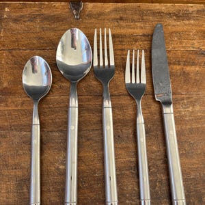 May include: A set of stainless steel silverware including a spoon, a large spoon, two forks, and a knife. The silverware has a simple design with a silver handle and a small, round accent near the top of the handle.
