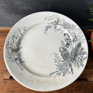 May include: Off-white ceramic plate with a floral design in shades of gray and black. The plate features a detailed botanical pattern of flowers, leaves, and grasses around the rim. The plate has a slightly aged appearance.