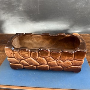 May include: A rectangular brown ceramic planter with a textured, stone-like exterior. The planter has a glossy finish and a smooth interior. It sits on a blue book on a wooden surface. A decorative item for plants.