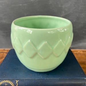 May include: A light green ceramic planter with a diamond pattern around the exterior. The small, round planter sits atop a stack of blue books. The interior of the planter is a matching light green color.