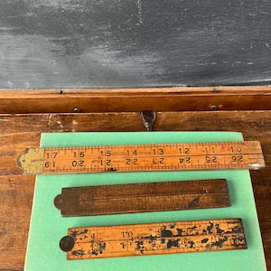 May include: Three vintage wooden folding rulers, each with inch measurements. The top ruler is open, showing numbers from 10 to 19 inches. The middle ruler is folded. The bottom ruler is partially open, showing numbers from 4 to 10 inches. The rulers are on a green surface.