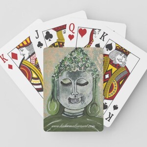 May include: Playing cards featuring a Buddha design on the card backs. The Buddha is depicted in green and grey, with a peaceful expression. The website address www.kabeemalewisart.com is printed on the card.