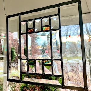 May include: Geometric stained glass window with a black metal frame. The design features clear and colored glass pieces in rectangular and square shapes. The window is hanging in front of a blurred outdoor scene.