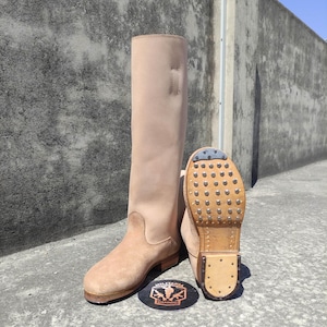 Pode incluir: Um par de botas de montar em couro castanho claro. Uma bota está em pé, a outra está angulada para mostrar a sola com tachas. As botas têm uma haste alta e uma biqueira arredondada. Um autocolante com o logótipo está na base das botas.