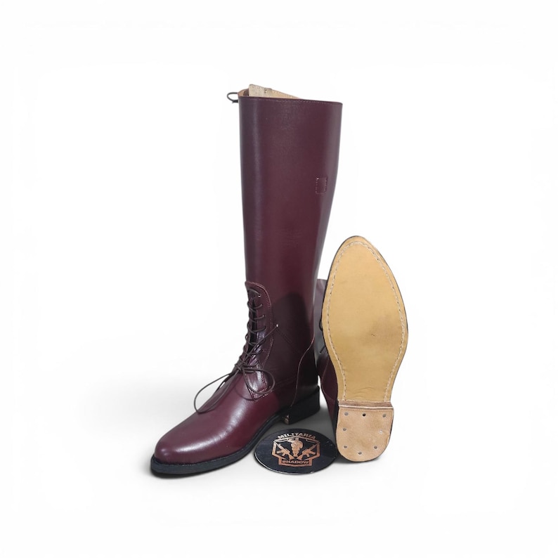 May include: Burgundy leather riding boots with a lace-up front and a smooth finish. The boots are tall, reaching the knee, and are displayed with a boot sole and a round emblem.