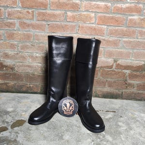 May include: A pair of tall, black leather boots with a shiny finish. The boots have a knee-high design and a round toe. A round emblem with the text "MILITARIA SHADOW" is placed between the boots.