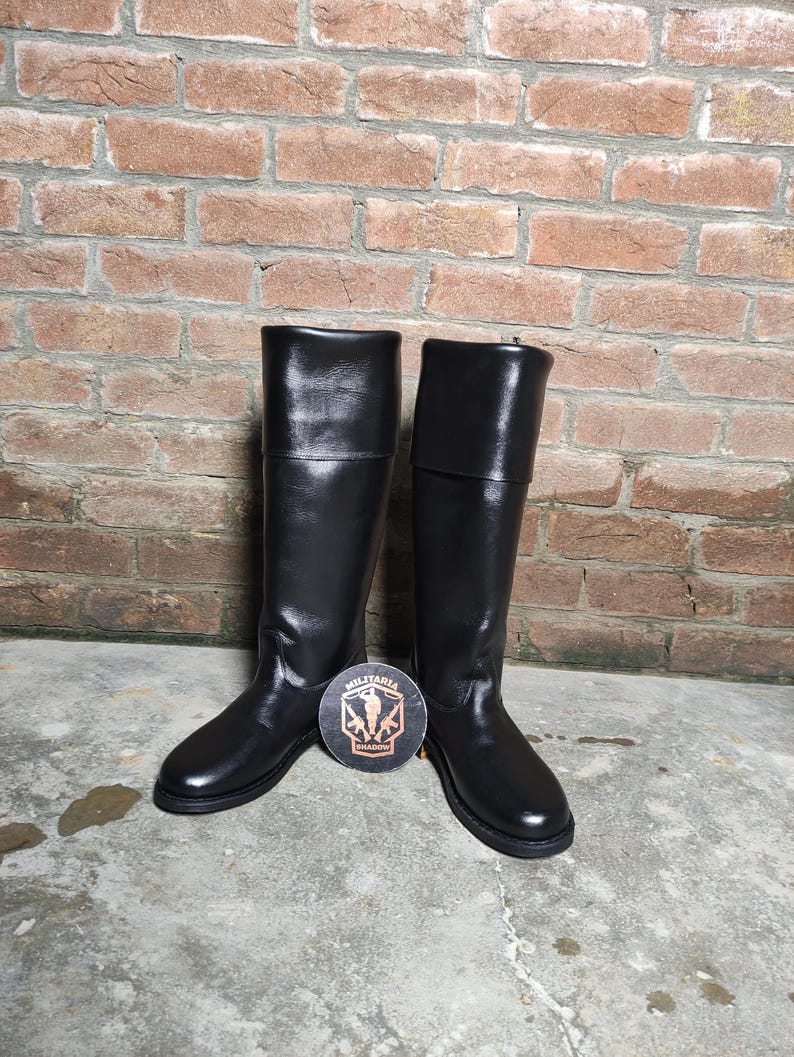 May include: A pair of tall, black leather boots with a shiny finish. The boots have a knee-high design and a round toe. A round emblem with the text "MILITARIA SHADOW" is placed between the boots.