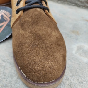 May include: Close-up of a brown suede and leather boot. The boot features a suede toe and a leather upper with black laces. The sole is stitched with white thread. The boot is a neutral color, suitable for various outfits.