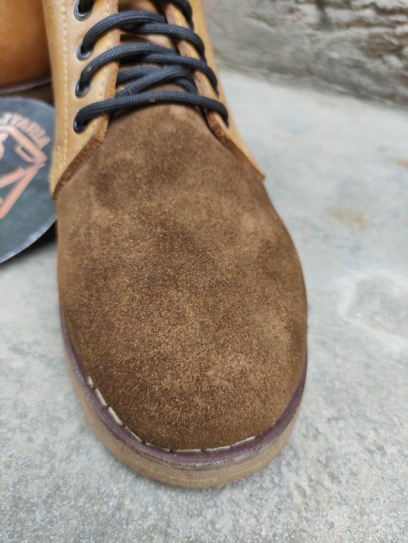 May include: Close-up of a brown suede and leather boot. The boot features a suede toe and a leather upper with black laces. The sole is stitched with white thread. The boot is a neutral color, suitable for various outfits.
