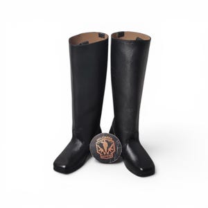 May include: A pair of black leather riding boots with a square toe and tall shaft. The boots are displayed on a white background with a round logo between them. The logo has the word "Militaria" on it.