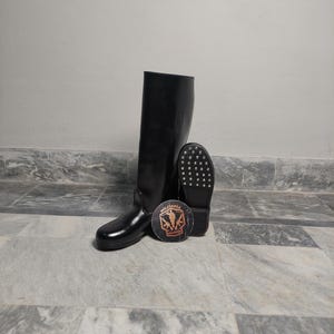 Puede incluir: Bota alta de goma negra con suela a juego y un parche redondo con la palabra "Militaria" y un gráfico de soldado. La bota está de pie junto a su suela. La bota está sobre una superficie de mármol.