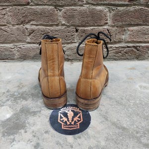 May include: A pair of tan leather boots with black laces, viewed from the back. The boots have a worn appearance and are set against a brick wall background. A round sticker with the words "MILITARIA SHADOW" is on the ground.
